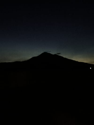 The image is a low-light, vertical, full shot of a silhouetted mountain range against a dark sky. The mountain's outline is prominent, with a slightly peaked summit. A small, faint white light is visible to the right of the mountain. The sky transitions from a deep, dark blue at the top to a lighter, muted gray and yellow near the horizon behind the mountains. The foreground is entirely black, obscuring any details of the terrain. The scene appears to be a natural landscape, possibly captured during twilight or at night. Without further context, the specific location in Keureuweungkrueng, Indonesia cannot be determined from the image alone.