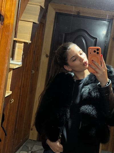 A woman is taking a selfie in a mirror. She is wearing a black jacket with a fur collar. The mirror reflects a black door. The background includes a wooden surface with trim. The location is unknown and cannot be determined based on the visual data.