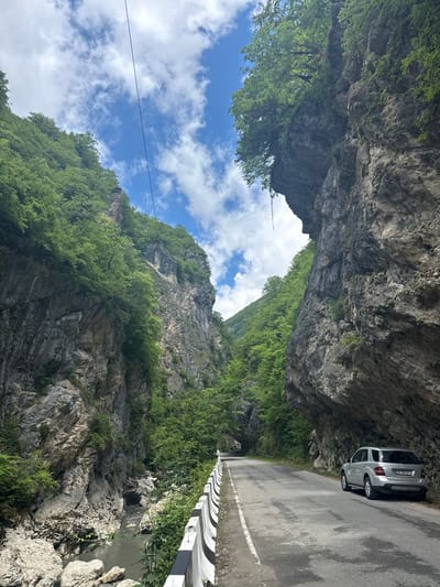 The image shows a silver Mercedes-Benz SUV parked on the side of a paved road that winds through a narrow gorge. The road is bordered by a concrete barrier with black and white markings. Steep, rocky cliffs rise on either side of the road, covered in lush green trees and vegetation. A river flows through the bottom of the gorge, between large rocks and boulders. The sky above is blue with scattered white clouds, and overhead wires are visible. The overall scene depicts a natural landscape in Achara, Georgia, likely a scenic route or a mountain pass.