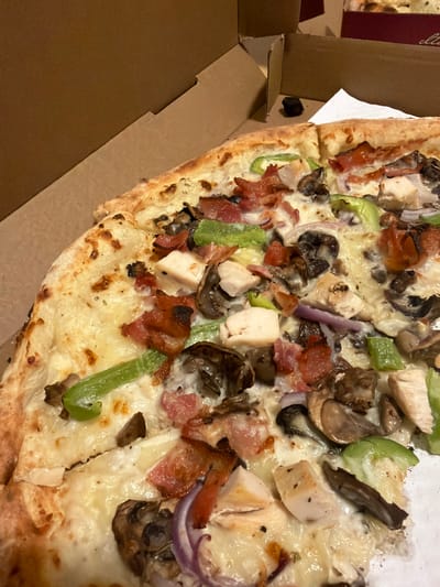 The image shows a close-up, overhead view of a pizza with various toppings, including chicken, bacon, mushrooms, green peppers, and red onions, all covered in melted cheese. The pizza is partially enclosed in a cardboard pizza box. The background is blurred, but appears to be the interior of a room with brown cardboard elements, suggesting a delivery or takeout setting. There are no people visible in the image, and no specific location in Edmonton, Canada is identifiable from the visual information provided. The scene is static and focuses solely on the food item.