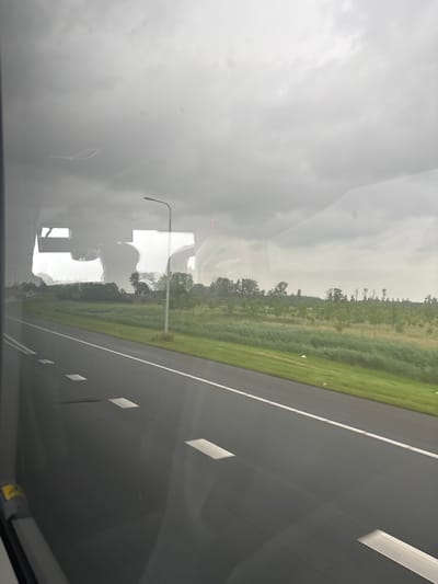 The image, viewed after a 90-degree counter-clockwise rotation, captures a highway scene from inside a moving vehicle. The right side of the frame features a multi-lane asphalt highway with white dashed and solid lane markings. To the left of the highway, a green, grassy expanse gives way to a line of scattered trees and dense foliage stretching towards the horizon. The sky, occupying the upper portion of the image, is overcast and hazy, indicating a cloudy or foggy daytime atmosphere.

The primary subjects are the highway, the natural landscape of grass and trees, and the hazy sky. Strong reflections on the vehicle's window are a notable element, obscuring parts of the external view. A large, bright, irregularly shaped reflection dominates the left-central area, possibly from an interior light source or a highly reflective object outside. A distinct reflection of a streetlight with a curved arm is visible near the center-left. Faint, abstract reflections, potentially from the vehicle's dashboard or internal structure, are also present, particularly in the lower left. Small, circular smudges or dust particles are visible on the window surface.

Faint red digital characters, appearing to be "E" and possibly "3", are barely discernible in the upper right quadrant of the reflected view. A small, yellow rectangular object with dark markings is faintly reflected in the lower left.

The image depicts an ordinary moment of travel on a highway through a rural or semi-rural setting. Given the provided context, the scene is located near Beesd, Netherlands, under overcast and hazy weather conditions during the day. No specific activity or emotional tone is explicitly conveyed beyond observation of the journey.