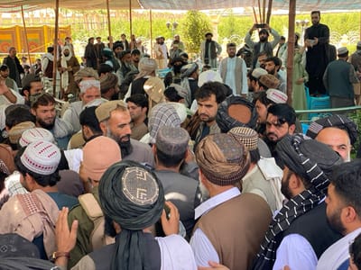 The image shows a crowd of men gathered under a white tent in the city of Zaker-E Sharif, Afghanistan, at the location of latitude: 31.547, longitude: 65.761. The men are all wearing traditional clothing, including turbans and long shirts. Many of them are standing, and some are sitting. The image is taken from a low angle, and the men's faces are visible. Some of the men are looking at the camera.  The men are standing in a crowded cluster.  The environment includes a white tent with fabric panels, a wooden bench in the background, green foliage, and several trees in the distance. The men are seemingly observing an event or awaiting something.