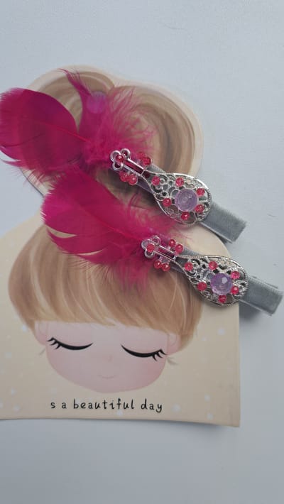 The image displays two decorative hair clips, each adorned with a silver filigree setting, pink rhinestones, and a larger purple gem. A cluster of delicate pink beads surrounds a central pink baguette-shaped stone on each clip. Attached to the bottom of each setting are two bright fuchsia feathers. These hair clips are placed on top of a notebook featuring a cartoon illustration of a young girl with blonde hair and closed eyes, her face tilted slightly. The text "s a beautiful day" is written vertically along the right side of the notebook in a simple, black font. The background is a plain, light-colored surface, possibly a desk or table. The overall impression is of a charming and whimsical accessory. The location context of Altai, Kazakhstan, does not provide specific visual cues within the image itself. There is no indication of the time of day or weather. The emotion conveyed is cheerful and innocent.