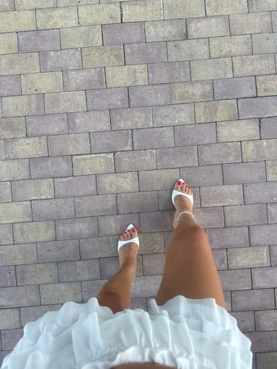 A person's legs are shown from a top-down perspective, walking on a paved walkway. The walkway is made of rectangular gray and beige bricks laid in a running bond pattern. The person is wearing white open-toed heels with a red pedicure and a silver anklet. A white ruffled skirt is visible at the bottom of the frame. The scene appears to be outdoors, with natural lighting.