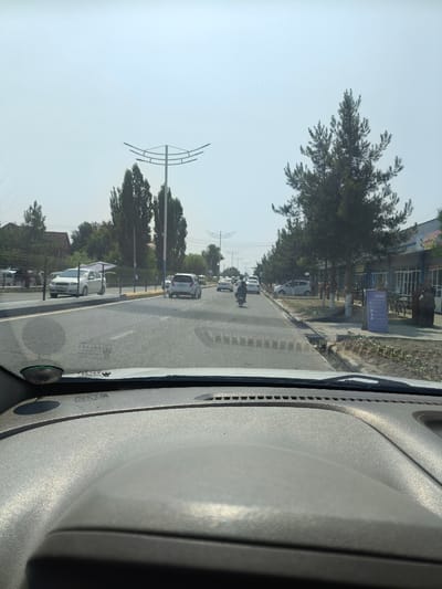 A perspective view from inside a vehicle shows a street scene in Paytugh, Uzbekistan, on a hazy, sunny day. The view is looking forward through the windshield. The street is paved, with multiple lanes of traffic. Cars and a motorcycle are visible on the road. On the right side of the road, there are buildings with storefronts, and people are visible standing outside. Trees line the sidewalk. On the left side of the road, there are more trees and some residential buildings. The sky is a pale blue with some haze. The dashboard of the vehicle is visible in the foreground.
