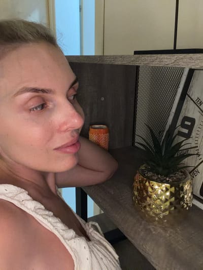 The image shows a close-up profile of a blonde woman with fair skin, looking to her left. Her hair is pulled back, and she is wearing a white, textured garment, possibly a top or dress. Her skin appears to have some redness around the nose and cheeks. In the background, a shelf holds several items: an orange patterned cylindrical object, a small potted plant with green leaves in a textured gold pot, and a decorative clock or sign with numbers and text, including "TEN," "NINE," "EIGHT," and "VEN." The environment appears to be indoors, with a hint of a doorway or partition in the upper left corner, suggesting a room setting. There is no indication of the location in Mojanovići, Montenegro.