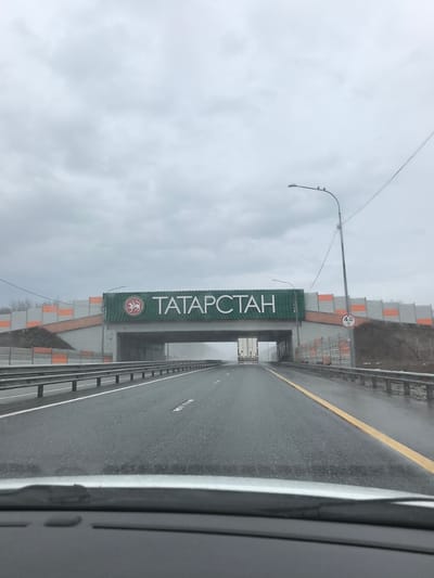 The image depicts a view from inside a vehicle, likely on a highway, approaching a bridge. The bridge is adorned with a large sign that reads "TATAPCTAH" (TATARSTAN in Russian), suggesting this is a border or landmark denoting entry into the Tatarstan region. The bridge structure is concrete with light gray and orange noise barriers on either side. A highway overpass sign on the right is marked "4.0 M" indicating a height restriction.

The road surface appears wet, possibly from rain. Highway guardrails are visible on both sides of the road. A truck is visible driving on the opposite side of the highway, passing under the bridge. The sky is overcast and gray. The location appears to be along a highway near Staroe Tyaberdino, Russia.