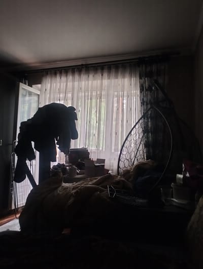 The image depicts the interior of a room, likely a bedroom, with soft natural light filtering through sheer curtains. The room appears somewhat cluttered. In the foreground, a bed is covered with blankets and appears unmade. To the left, a dark silhouette of a stand holding several pieces of clothing is visible against the window. Beyond the bed, a hanging chair with a woven design is partially visible. Further to the right, on a surface that could be a bedside table or dresser, are various objects including stacked items, containers, and a glass. The overall impression is a lived-in space, with shadows obscuring some details due to the lighting conditions. There are no visible people in the image. The specific location in Zyuzino, Russia, cannot be determined from the visual information provided.