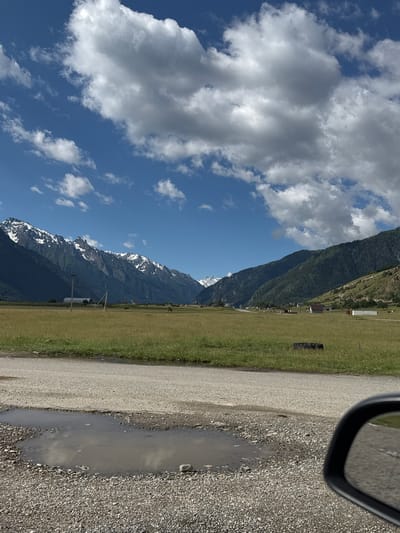 The image captures a scenic mountain landscape under a partly cloudy sky. The foreground features a gravel road with a puddle of water, reflecting the sky and surrounding elements. To the left of the road, a vast grassy field stretches out, dotted with a few scattered structures that appear to be farm buildings or sheds. Beyond the field, a majestic mountain range dominates the background. The lower slopes of the mountains are covered in dense green vegetation, while the higher peaks are dusted with snow, indicating a cooler climate or higher altitude. The sky is a vibrant blue, interspersed with large, fluffy white clouds. The sun appears to be shining, casting shadows and highlighting the textures of the landscape, suggesting it's likely daytime. The overall mood is serene and picturesque, evoking a sense of natural beauty and tranquility. The image was taken from a vehicle, as suggested by the presence of a car's side mirror in the upper right corner. No visible text is present in the image. The location context places this scene in the Verkhne-Teberdinskoe rural settlement in Russia.