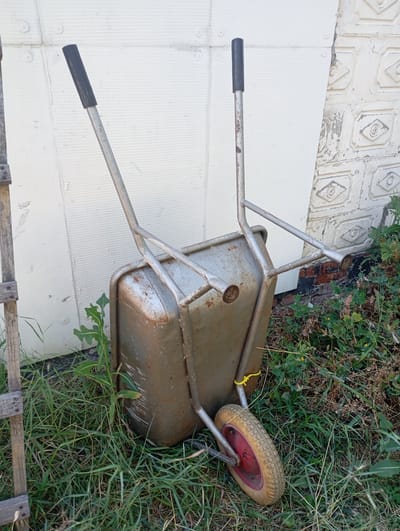 A weathered, single-wheeled wheelbarrow rests on a patch of grass in front of a white, textured wall and a brick wall with decorative elements. The wheelbarrow is made of metal and shows signs of rust and use. It has two silver handles with black grips, and a red-rimmed, yellow tire. A yellow tie is wrapped around one of the wheelbarrow's handles. Green weeds and grass surround the wheelbarrow, with some dry foliage visible behind it. To the left of the wheelbarrow, a wooden ladder or fence post is partially visible. The scene appears to be outdoors, possibly in a yard or garden, during daylight hours. There is no discernible text in the image. The setting is Skotuvate, Ukraine.