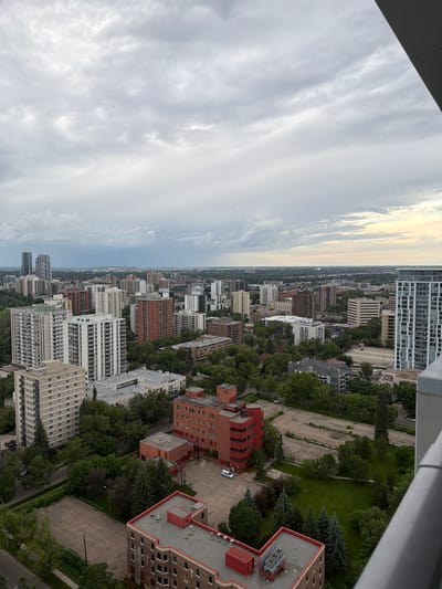 The image is an aerial view of a city skyline on a cloudy day, likely taken from a high-rise building in Edmonton, Canada. The foreground features the corner of a balcony railing. Below, the scene unfolds with a dense urban landscape characterized by numerous multi-story apartment buildings and residential complexes. Greenery, in the form of trees and parks, is interspersed throughout the cityscape, suggesting a well-treed urban environment. In the mid-ground, several distinct buildings are visible, including a tall, reddish-orange, spiral-shaped structure and a long, lighter-colored apartment building. Further in the distance, more buildings of varying heights and architectural styles stretch towards the horizon, merging with the overcast sky. The overall impression is one of a sprawling, developed urban area with a noticeable presence of residential housing and green spaces.