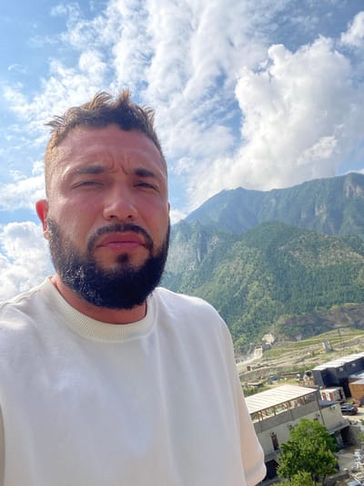 The image is a selfie taken by a man outdoors, with a mountain landscape in the background. The man is facing the camera, with his head and shoulders visible. He has short, spiky brown hair, a full beard, and is wearing a light-colored crew neck sweatshirt. His expression is serious. Behind him, lush green mountains rise up, with a clear blue sky dotted with white clouds. In the mid-ground, a town or village is visible, with buildings, roads, and what appears to be a hydroelectric dam or construction site. The style of the buildings suggests a rural or mountainous region, and the presence of the dam indicates potential industrial activity.
The location is identified as Chalda, Russia. This context suggests that the mountain range is part of the Caucasus Mountains. The presence of the dam and other structures could be related to infrastructure development or energy production in the region. The overall scene depicts a man in a scenic, somewhat remote, mountainous environment.