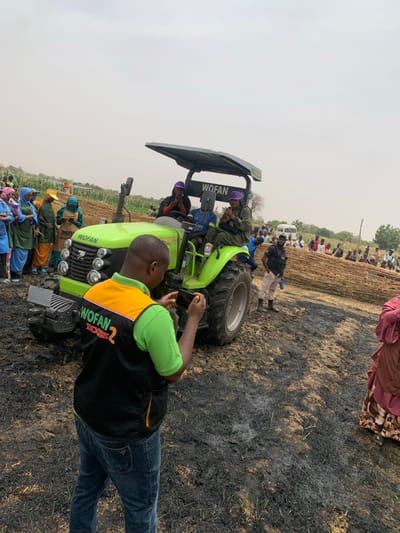 The image shows a green tractor in a field with burned vegetation. The tractor has a black canopy with the word "WOFAN" written on it. Three people are sitting on the tractor. A man in a green vest with the words "WOFAN ICON 2" printed on it is standing in front of the tractor and taking a picture with a smartphone. There are many people standing behind the tractor. The image was taken in Ringimawa, Nigeria (latitude: 11.666, longitude: 8.414). The field has patches of burnt vegetation. A white vehicle is visible in the background.