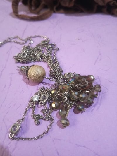 A metallic chain necklace with multiple decorative elements is displayed on a textured, light purple surface. The necklace features a prominent spherical bead with a granulated, dull gold or brass-like finish, approximately 1.5 cm in diameter. Adjacent to this, a cluster of numerous small, faceted, iridescent beads, appearing brownish-grey with subtle purple and green reflections, are attached. The chain itself is silver-colored and includes intermittent square-shaped links, some of which contain small, clear, brilliant-cut stones. A section of a dark brown, possibly fabric, object is visible out of focus in the upper background. Several strands of dark hair are present near the cluster of iridescent beads. The overall arrangement of the necklace suggests it is casually laid out or slightly tangled. The scene is located in Chandragiri, Nepal.