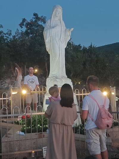 The image depicts a gathering at Apparition Hill in Međugorje, Bosnia and Herzegovina, at dusk. Dominating the scene is a large, white statue of the Virgin Mary, holding a rosary. In the foreground, a family is present: a man in a white t-shirt and beige shorts, a woman in a beige dress holding a toddler, and another man in a white t-shirt and beige cargo shorts, wearing a pink backpack with the name "Kiara" visible. The area is cordoned off by a metal railing, with decorative plants and flowers visible behind it. Several lit candles and a sign with Croatian text, possibly indicating "Attention," are also present. The background consists of trees and a hillside under a twilight sky.
