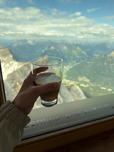 A hand holds a glass of coffee in front of a panoramic view of mountains. The coffee appears to be an espresso with foamed milk. The hand is of a person wearing a beige jacket with gathered elastic at the cuff. The background shows a vast landscape of green valleys and rocky mountain peaks under a cloudy blue sky. The image is taken from inside a building, with a window framing the view. Text is visible on the window, suggesting a location in Ehrwald, Austria, possibly related to a cable car or viewpoint, as the words "Ehrwald" and "Silvretta" are partially legible. The lighting suggests it is daytime.