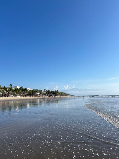 A beach scene under a clear, bright blue sky. The shore is lined with palm trees and buildings. A long pier extends into the water. Gentle waves with white foam roll onto the sandy beach. Mountains are visible in the distance, partially obscured by clouds. The water reflects the sky and surrounding environment, creating a mirrored effect on the wet sand.