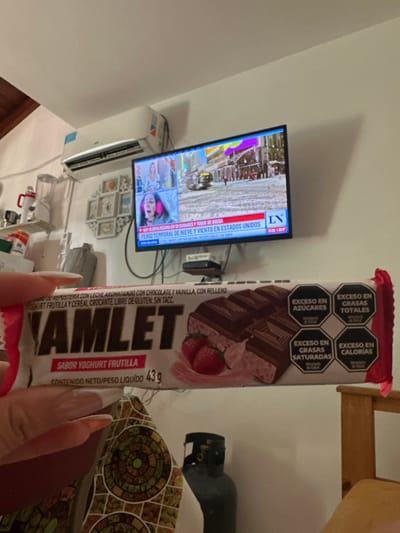 The image shows a Hamlett yogurt strawberry-flavored chocolate bar with a warning label indicating it is high in sugar, saturated fat, total fat, and calories. In the background, a television displays a news report about severe snow and windstorms in the United States. Also visible are parts of a room, including an air conditioner, decorations, and a chair. A hand with long fingernails is holding the candy bar.
