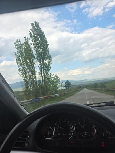 A panoramic view captured from the interior of a vehicle, looking out through the windshield, is presented. The image is oriented with the horizon running vertically, suggesting it was taken with a rotated device.
In the foreground, the dark grey steering wheel and a section of the dashboard are visible on the right side. The instrument cluster shows a speedometer with readings up to "240 km/h", currently resting at "0 km/h". A tachometer reads "x1000 r/min" and also shows zero. A digital display indicates "288339" (likely an odometer reading) and "11:33" (the time). A red warning light, possibly for brakes or another system, is illuminated on the top right of the instrument cluster. An air vent is also visible on the dashboard.
Outside the vehicle, a narrow asphalt road stretches into the distance, framed by green grassy verges. Tall, slender trees with lush green foliage, possibly poplars, line the road, particularly prominent on the left side of the frame. Beyond the immediate landscape, rolling hills and mountains create a layered backdrop under a partially cloudy, blue sky. A body of water, possibly a lake or river, can be glimpsed in the mid-distance between the trees and the hills.
The scene depicts a peaceful daytime setting, with the vehicle seemingly stationary, allowing for observation of the scenic environment. The time displayed, 11:33, confirms it is late morning. The location is contextualized as Lelintsi, Bulgaria, suggesting this natural landscape is part of the region's scenery. A sticker with illegible text is affixed to the right side of the windshield.