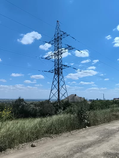 A tall, lattice steel power pylon stands centrally in a landscape under a bright blue sky with scattered white clouds. The pylon is surrounded by trees and vegetation, with a dirt road in the foreground. In the distance, a town or city is visible, with buildings and what appears to be a smokestack or industrial structure. The scene appears to be located in or near Varnița, Moldova.