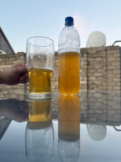 A glass of beer and a plastic bottle of beer are on a reflective surface outdoors. The beer in the glass is about halfway full and has a foam head. The bottle is full of beer and has condensation on its exterior. In the background, a brick wall and a clear sky are visible. A person's hand is partially visible in the left side of the frame. The scene appears to be on a rooftop in Khazar Raion, Azerbaijan.