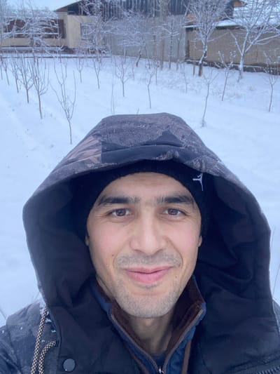 A man wearing a hooded jacket stands outside in the snow. A building is visible in the background along with small trees covered in snow. The man is wearing a black beanie beneath the hood, and a light blue jacket underneath the outer layer. His face is turned towards the viewer with a slight smile. The jacket's hood is grey with darker patterns. The snow-covered ground is bright.