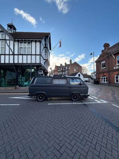 The image shows a grey Volkswagen van parked on a brick-paved road with white parking lines in Berkhamsted, Dacorum, United Kingdom. The van is situated next to a black and white Tudor-style building with multiple windows and a Union Jack flag flying nearby. Across the street are brick buildings of varying heights. The sky is blue with scattered clouds, suggesting a sunny day.