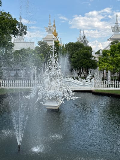 A white ornate fountain stands in a dark pond in front of a complex of white buildings. The fountain features intricate, swirling designs resembling mythical creatures and lotus flowers. Water sprays upwards and outwards from the fountain, creating a misty veil. In the background, the white buildings exhibit distinctive Thai architectural features with tiered roofs and pointed spires. Lush green trees frame the scene, and a bright blue sky with scattered white clouds provides the backdrop. The location is identified as the White Temple (Wat Rong Khun) in Ban Rong Khun, Thailand. There are no people visible in the image.
