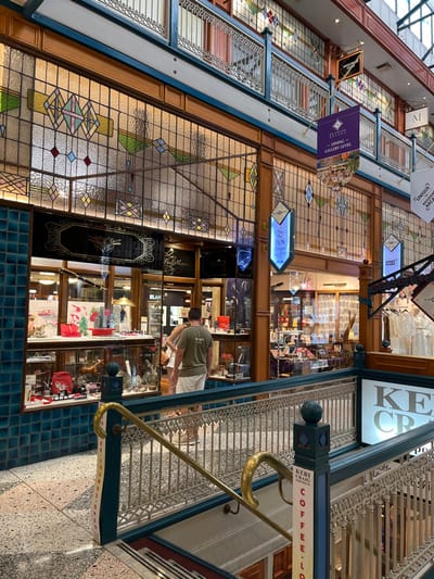 This media file captures the interior of a multi-level shopping arcade, presenting a blend of historic architecture and contemporary commerce. The setting is characterized by elaborate stained glass panels that form parts of walls and shopfronts, featuring geometric patterns and subtle colors. Ornate metal balustrades with intricate scrollwork line the upper levels, complementing gold-colored handrails that guide visitors.

Two individuals are visible in the lower right (when oriented correctly), looking into a shop display. One person wears a green t-shirt and white shorts. The shops feature polished wooden frames and glass showcases, displaying a variety of goods, including what appears to be luxury pens (part of "BLAN", likely Montblanc, is visible) and other gift items. The flooring is a mix of patterned blue tiles and speckled terrazzo.

Prominent signage indicates various establishments and levels. Text visible includes "DINING," "GALLERY LEVEL," "M," "KERI CRAIG COFFEE-LO," and fragmented words like "UNIQUE," "BRISBANE," and "ARCA," strongly suggesting the location is a historic arcade in Brisbane. The overall lighting suggests it is daytime, with light filtering through the upper structure or internal lighting. The scene portrays a calm, everyday retail environment within a beautifully preserved architectural space.
