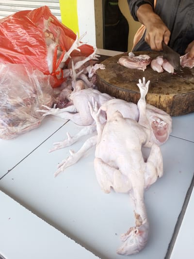 In Nhot Chang, Laos, a butcher cuts up raw chickens on a wooden board. Several whole, defeathered chickens are arranged on a white tiled surface, and a clear plastic bag and a red plastic bag contain chicken parts. The butcher, wearing a grey long-sleeved shirt, uses a large cleaver to cut the meat. The scene is a market setting with yellowish walls in the background.