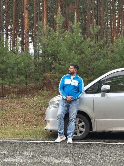 A man is leaning against a silver car parked alongside a road next to a forest. The man is wearing a blue Adidas track jacket, light-wash jeans, and white sneakers with orange stripes. The car is parked very close to the edge of the road. The road is paved and has a white line marking the edge. The forest consists of tall pine trees. The photograph was taken in Сизая, Russia.