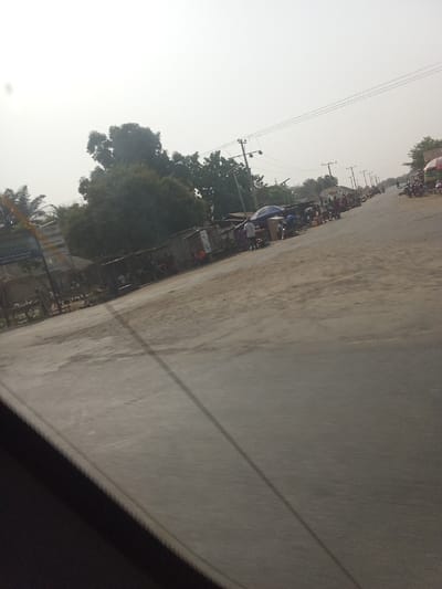 This image depicts a roadside market scene in Marke, Nigeria, viewed from inside a vehicle. A paved gray road occupies the center and foreground, flanked by unpaved, dusty shoulders. In the lower-left foreground, a dark, angled structure suggests a vehicle's interior.
Along the left side of the road, a series of makeshift market stalls, constructed from corrugated metal sheets and wood, are visible. Various goods are displayed on and around these structures. Several individuals, including adults and possibly children, are standing or milling about near these stalls. Dense green trees and foliage are present behind these structures, with a partially visible blue and white sign on the far left.
Further down the road, on the right side, market activity continues with more stalls, people, and several motorcycles parked or in motion. Two prominent open umbrellas, one blue and one with multiple colors, provide shade over market areas. A series of wooden utility poles carrying overhead power lines run parallel to the road, receding into the distance. The background shows the road extending with more distant structures and vegetation under a bright, hazy sky.