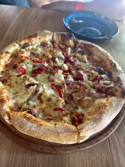 A close-up, overhead shot captures a whole pizza, sliced into eight pieces, resting on a round, wooden pizza board. The pizza, with a thick, golden-brown crust that's slightly charred in places, is generously topped with melted cheese, pieces of chicken, and sliced red peppers and possibly sausage. The toppings appear glistening and freshly cooked.
To the left of the pizza, a dark, shallow bowl is partially visible. A person's arm, wearing a red wristband, is also seen in the frame, suggesting they are the one about to serve or eat the pizza. The background consists of a light-colored wooden table, indicating an indoor setting, possibly a restaurant or home dining area. The lighting is bright, suggesting daytime.
The scene evokes a sense of casual dining and enjoyment. There is no visible text in the image. The overall impression is of a delicious, ready-to-eat pizza on a rustic wooden surface, with a hint of human presence.