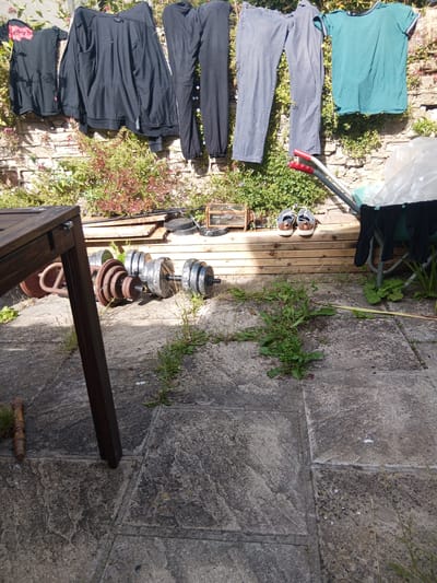 A collection of clothing is hung on a washing line against a stone wall. Among the clothing items are black long-sleeved tops, black trousers, and a teal t-shirt. The washing line is also adorned with two pairs of shoes, one pair grey and brown, the other grey and white. On the ground in front of the washing line are several objects, including two weights, a brown wooden chair leg, and a wooden plank structure. The scene appears to be an outdoor patio area with flagstone flooring. The image does not provide enough information to determine the specific location in Bristol, United Kingdom.