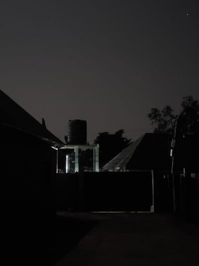 The image depicts a nighttime scene in Kason, Nigeria. In the foreground, a dark, angled structure, likely a roof or wall of a building, dominates the left side of the frame. To the right of this, a large, cylindrical water tank stands atop a lighter structure, possibly a small building or platform. This tank appears to be a prominent object in the scene. Further to the right, a building with a distinctive sloped roof is visible. The background features trees silhouetted against the dark night sky. A faint light source, potentially a distant star or light, is visible in the upper right portion of the sky. The overall impression is one of a residential or industrial area at night, with functional infrastructure like water storage being central to the composition.