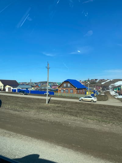 The image shows a street in Verkhniye Tatyshly, Russia. The view is from a moving vehicle. A bright blue sky occupies the left portion of the frame. On the right side of the road, there is a house with a distinctive blue roof and yellow and blue vertical stripes along its sides. A white car is parked adjacent to the house. Further up the road, a village or small town landscape extends into the distance. Utility poles and power lines run alongside the road. Patches of snow are visible on the distant hills. The roadside is earthen and grassy. The road itself is asphalt.