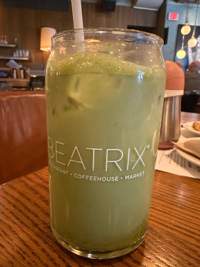 The image shows a partially consumed green-colored beverage in a glass bearing the logo "BEATRIX RESTAURANT • COFFEEHOUSE • MARKET." A white straw is inserted into the drink. The glass sits on a wooden table with visible grain patterns. The background reveals an interior setting, possibly a restaurant or cafe, with other tables, chairs, and some lighting fixtures visible. It is inferred that the image was taken at or in proximity to the Beatrix restaurant in Oak Brook, Illinois, United States, based on the logo on the glass. No people are clearly visible in the foreground, but blurred figures can be seen in the background. The general environment suggests a casual dining or coffee shop atmosphere.