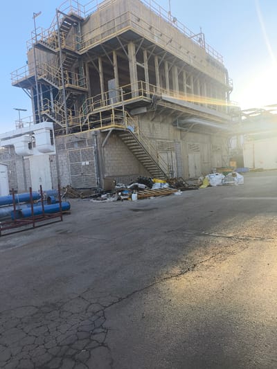 This is a medium shot, outdoor, eye-level photograph of an industrial facility with a large concrete and metal structure under a clear blue sky. The structure appears to be a processing plant or a storage facility, with multiple levels, staircases, and railings. The building is made of concrete blocks and metal beams, with some sections having a weathered, industrial aesthetic. Yellow railings and staircases are prominent features of the building's exterior.
In the foreground, there is a paved area, likely a yard or driveway, showing some cracks and wear. To the left, several large blue pipes are stacked on wooden pallets and secured by a red metal frame. Further into the scene, near the base of the structure, is a collection of debris and waste. This includes several large white bags, some yellow bags, wooden pallets, scattered trash, and a propane tank. The sun is shining brightly, casting lens flare into the image, particularly on the right side, suggesting it is either early morning or late afternoon. The overall impression is one of an operational industrial site, possibly undergoing maintenance or in a state of disarray due to waste management.
This photograph was taken in the city of Dimona, Israel.