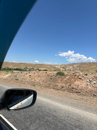 The image depicts a view from a car traveling through a rural area, likely on a sunny day. In the foreground, the asphalt road is visible, with the side mirror of the car on the left. The mirror reflects a person in a blue and white patterned garment. The exterior of the mirror housing shows signs of rust. Beyond the road, the landscape consists of dry, brown hills dotted with small buildings and some greenery, suggesting a village or settlement. The sky is a clear blue with a few scattered white clouds. The overall environment appears arid and mountainous. Based on the provided location information, this scene is situated in the city of Беш-Мойнок, Kyrgyzstan.
