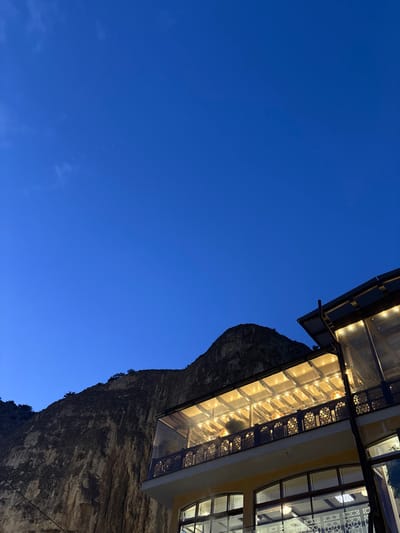 The image depicts a nighttime or late dusk scene in Bakhchysarai, Ukraine, featuring a multi-story building against a mountainous backdrop under a deep indigo sky. A rugged, dark rock formation or cliff face occupies the left and central background, with sparse vegetation on its upper edges. The building, situated in the lower right, prominently displays an illuminated upper-level balcony or terrace. This area is brightly lit by numerous warm-toned overhead lights, visible through a transparent or translucent roof structure and reflecting off clear enclosure panels. An intricately patterned dark balustrade with geometric or organic designs borders the balcony. The lower section of the building features large windows, some with decorative white grilles. No individuals, specific actions, or interactions are visible. The environment is stationary, characterized by artificial light against the natural dark terrain and twilight sky.