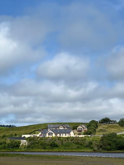 A wide, bright, daytime, full-frame, vertical shot depicts a scenic view of several modern houses situated on a grassy hill overlooking a body of water. The sky is predominantly blue with scattered, puffy, white and grey clouds. The houses are clustered together, with varying architectural styles, featuring light-colored walls, dark roofs, and numerous windows. Some of the houses have stone accents. The hill is covered in lush green grass, with scattered trees and shrubs. A white fence runs along the top of a section of the hill, and a road with a few cars is visible near the houses. In the foreground, there is a muddy, shallow body of water with a line of small, white birds on the shore. The overall impression is one of a tranquil, rural landscape in Tullycullion, Ireland, bathed in natural light.
