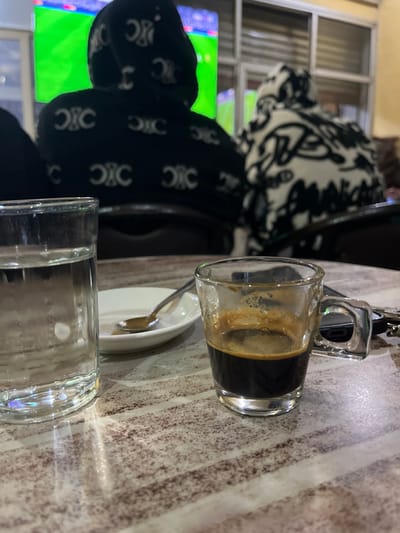 The image depicts an indoor scene within a cafe in Ouaouizeght, Morocco. In the foreground, a clear glass containing water is situated on the left, next to a small, white, oval saucer holding a silver spoon. To the right, a small clear glass cup contains dark espresso with a layer of crema. The table surface is light-colored with horizontal, irregular brown striations. A black smartphone and a set of keys are partially visible next to the espresso cup.

In the midground, two individuals are seated with their backs to the viewer. The person on the left wears a black hooded garment patterned with repeating white "C" or crescent-shaped logos. The person on the right wears a light-colored hooded garment with black, abstract, or graffiti-style patterns. Both individuals are oriented towards a large, brightly illuminated screen in the background.

The background features a large display screen showing a predominantly green image, suggesting a sports broadcast. Behind the screen, framed glass partitions or windows are visible, along with a light-colored wall. The environment suggests a public viewing area.