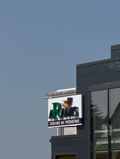 The image shows a close-up view of a sign for "ARAMS CENTRU DE PEDIATRIE" attached to a dark-colored building. The sign features green and black lettering, with a stylized orange and purple geometric shape above the text. The building appears to be modern, with a large window reflecting trees and sky. The sky is overcast and a pale blue-grey. The sign is illuminated by overhead lights, suggesting it is nighttime or the building is interiorly lit. The setting is consistent with an urban environment, likely a medical facility. Without further information or context, the precise location in Suceava, Romania cannot be determined from the image alone.
