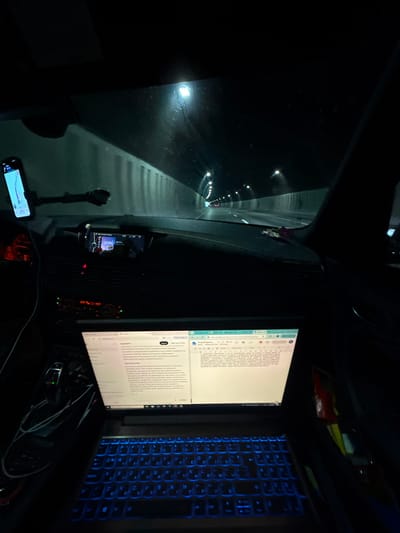 The image depicts the interior of a car driving through a tunnel at night. A laptop with a blue-backlit keyboard is open on the passenger seat, displaying a document. The laptop screen shows a Google Docs document, with text visible in a foreign language, likely Bulgarian. To the left of the laptop, a car's gear shift and center console are visible. A smartphone mounted on the dashboard displays a navigation app. The tunnel walls are lined with lights, and the road ahead is visible, illuminated by the car's headlights and tunnel lighting. The overall scene suggests someone is working or documenting while traveling through the tunnel. The location is identified as the tunnel on the road to Dolna Vasilitsa, Bulgaria.