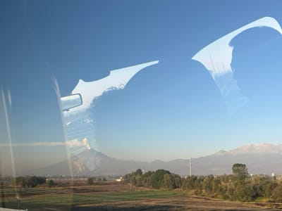The image captures a landscape view from within a vehicle, looking out across the Santa Ana Xalmimilulco region in Mexico, under a clear blue sky. Reflections on the vehicle's window include the back of a person's head with curly hair on the left, and the profile silhouette of a second individual, possibly the driver, with short hair, positioned to the right. A rearview mirror's reflection is also visible on the left.

The exterior environment features a prominent volcanic peak, identified as Popocatépetl, situated towards the left of the frame, emitting a distinct plume of smoke or ash. To the far right, a second mountain range, consistent with the snow-capped peaks of Iztaccíhuatl, rises on the horizon. The midground consists of agricultural fields or cultivated land, varying in color from green to brown, interspersed with trees and scattered urban or semi-urban structures. Utility poles are visible vertically in the midground. A faint horizontal line across the lower frame suggests a distant road or pathway traversing the fields.