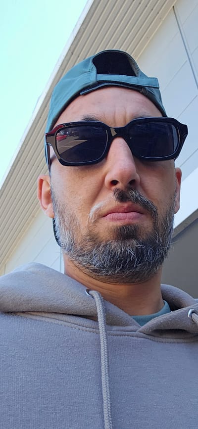 A close-up, low-angle portrait features a man with a salt-and-pepper beard and mustache, looking slightly upwards. He is wearing dark, rectangular sunglasses that cover his eyes. On his head, he wears a dark green baseball cap, positioned with the brim facing backward. He is dressed in a light grey hooded sweatshirt, the fabric of which shows some pilling, and a grey drawstring is visible across the front. A hint of a light blue garment can be seen under the collar of his hoodie.
The background consists of light-colored, horizontally paneled siding of a building on the left and a clear, bright blue sky in the upper right. The bright lighting suggests it is daytime with good weather. There is no visible text in the image. The man's expression is obscured by the sunglasses, and no specific activity is taking place other than posing for the photograph. The setting appears to be outdoors next to a building, possibly in Norwich, United Kingdom as per the provided context.