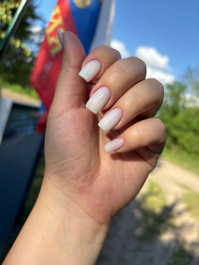 A close-up shot shows a left hand with white, glittery, square-shaped nails held up against a blurry background of a flag and foliage. The flag appears to be red and blue with some yellow emblem. The background suggests an outdoor setting on a sunny day, with a clear blue sky and scattered white clouds. The focus is on the hand and nails, with the background providing context but not detailed information about the location in Zubov, Russia, or specific actions.