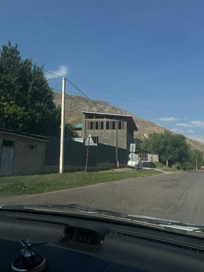 A residential street in Yakkatut, Uzbekistan, is depicted on a sunny day. The scene is viewed from inside a vehicle, looking forward through the windshield. On the left side of the road, there is a single-story brick building with a metal door and windows. Adjacent to it is a dark green metal fence. A tall utility pole stands in front of the fence, with electrical wires running across the frame. Beyond the fence, a large, multi-story building with a prominent upper floor featuring large, multi-pane windows is visible. The building appears to be under construction or in a state of renovation, with exposed rafters on the roof. In the background, a steep, arid hillside dominates the landscape. To the right of the road, there are trees and another smaller building, as well as a white car covered with a tarp. The road itself is paved and appears to be in moderate condition. The sky is clear blue with a few wispy clouds. The dashboard of the vehicle is visible in the foreground, with a decorative item on the left and a small electronic display on the right.