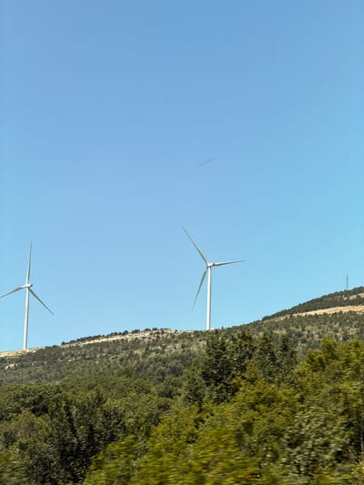 The image depicts a rural landscape with two wind turbines on a grassy hill under a clear blue sky. The wind turbines are white and have three blades each. The hill is covered in green vegetation, including trees and shrubs. In the distance, a single power pole is visible. The foreground is filled with lush green trees. The image appears to have been taken from a moving vehicle due to the motion blur in the foreground foliage. The scene suggests a location in a windy, open area, consistent with renewable energy generation. This specific scene is located in Bratica - Braticë, Montenegro.
