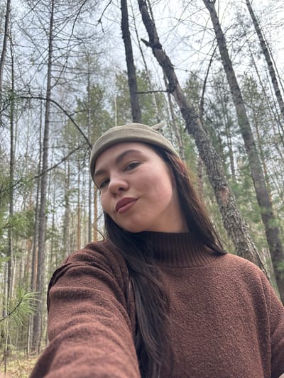 A person is depicted in a selfie format within a wooded area. The subject is wearing a brown, textured sweater and a light green cap. Their hair is dark and falls to the side. The background consists of a forest setting, with numerous trees exhibiting bare branches against an overcast sky. The location is near Kachkanar, Russia, as the trees appear similar to that of a birch forest. A single pine branch is visible in the lower center of the frame.